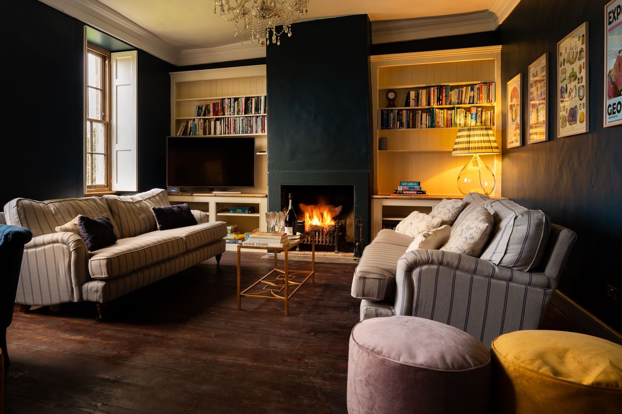 Drawing room with deep navy walls, a lit open fire, striped sofas and built-in bookshelves